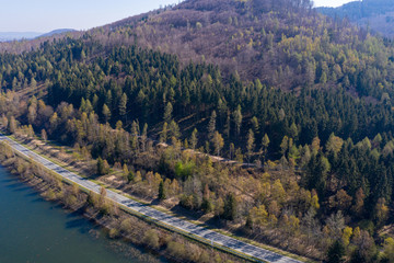 Aerial view of an asphalted country road along the edge of a densely wooded mountain on the bank of a dam.