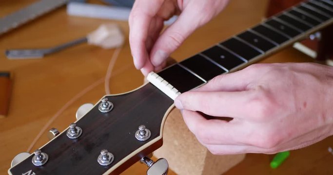 Close up hands of a luthier fitting and gluing a nut onto an acoustic guitar neck fretboard on a wood workbench with lutherie tools.