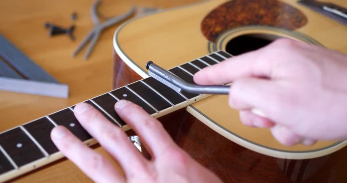 Close up hands of a luthier filing and crowning the frets of an acoustic guitar neck fretboard on a wood workbench with lutherie tools.