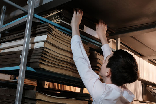A student in a white shirt pulls out an old book from the top bookshelf in the library or in the archive room. Schoolboy studying historical documents. Concept back to school or popularize reading.