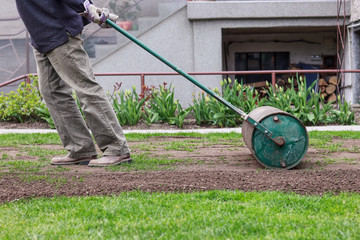 Man in working outfit pulls lawn roller behind. Necessity after long winter and spring for flat...