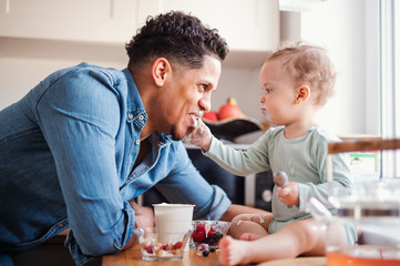 A father and a small toddler son eating fruit and yoghurt indoors at home.