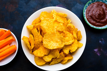 Crispy potato chips in a bowl with carrot slices and tomato sauce on a textured background.