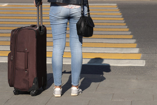 Woman with a suitcase on wheels stands on a pedestrian crossing, back view, shadow on road. Female legs in blue jeans on the crosswalk, concept of traveler, waiting taxi, street safety
