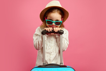 Smiling little girl in sunglasses and straw hat, holding suitcase on isolated pink, sincerely...