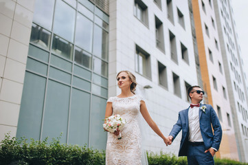 Stylish newlyweds stand holding hands in a modern city, against the backdrop of a high-rise building. Wedding portrait of fashionable groom in a blue suit and glasses and a bride in a white dress.