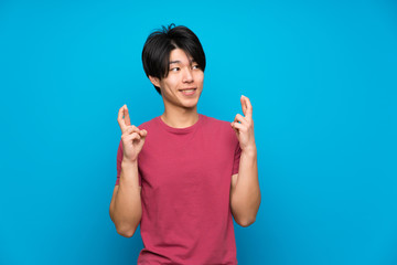 Asian man with red shirt over isolated blue wall with fingers crossing