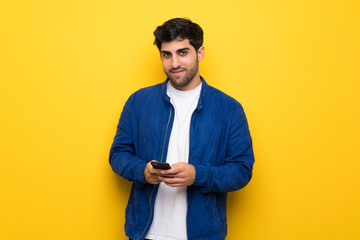 Man with blue jacket over yellow wall sending a message with the mobile