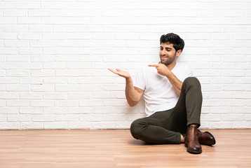 Young man sitting on the floor holding copyspace imaginary on the palm to insert an ad