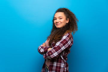 Teenager girl over blue wall keeping the arms crossed in lateral position while smiling