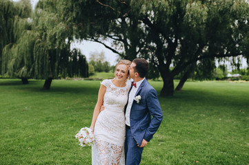 Young groom in a blue suit walking with a beautiful blonde bride in a park with green grass, trees and willows. Wedding portrait of smiling and loving newlyweds. Hot kiss.
