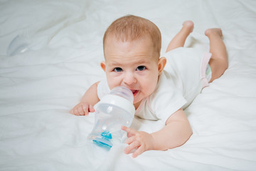 Infant baby with big brown eyes lying on the white bed with special bottle of water with nipple. Tries to gnaw her. teething