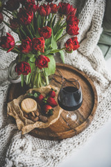Fototapeta na wymiar Wine snack set with flowers. Glass of red wine, cheese, roasted almonds, strawberries and bouquet of Spring red tulips on wooden tray over white knitted blanket, top view. Romantic mood concept