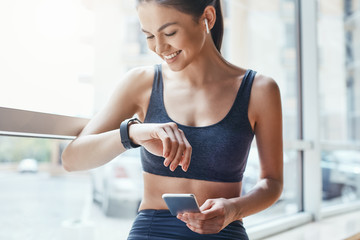 Making training easier. Young and cheerful woman in sportwear looking at her sport bracelet and using smartphone while standing in front of window at gym