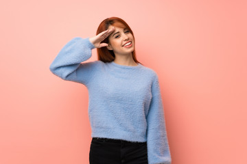 Young redhead woman over pink background saluting with hand