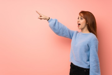 Young redhead woman over pink background pointing away