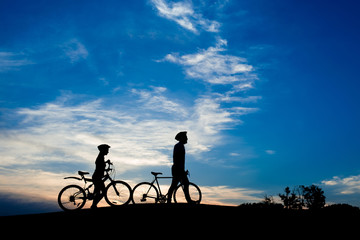 Silhouettes of young couple with bikes at sunset. Girlfriend with boyfriend walking with bicycles on evening sky background. Romantic date on bikes.