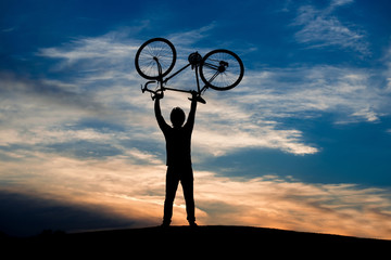 Silhouette of cyclist lifting bicycle at sunset. Silhouette of the man lifting a bike above his head on the meadow, evening sky background.