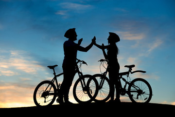 Cheerful couple of bicyclists clapping hands at sunset. Happy young couple having fun riding a bicycle on evening sky background.