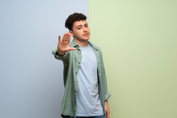 Young man over blue and green background making stop gesture denying a situation that thinks wrong