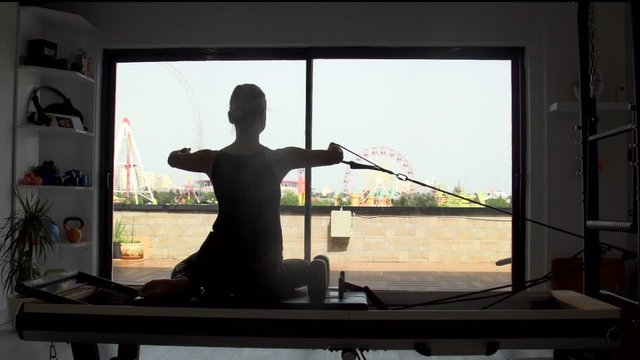 Silhouette of an athletic woman doing reformer pilates on the machine with backlight, amusement park scenery on the back