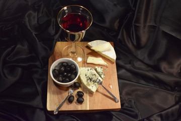 A glass of red wine, olives and two kinds of French cheese on a black background. French brie with white mold, cheese with blue mold. Soft brie cheese and blue cheese. Wood surface. 