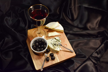 A glass of red wine, olives and two kinds of French cheese on a black background. French brie with white mold, cheese with blue mold. Soft brie cheese and blue cheese. Wood surface. 