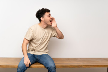 Young man sitting on table shouting with mouth wide open to the lateral