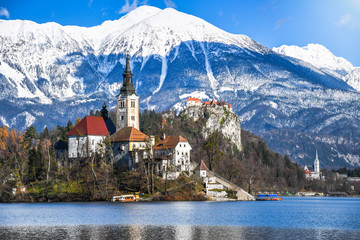Small natural island in the middle of alpine lake with church dedicated to assumption of Mary and castle with snowy mountain range in the background in winter landscape Bled, Slovenia - obrazy, fototapety, plakaty