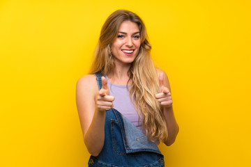 Young blonde woman with overalls over isolated yellow background points finger at you