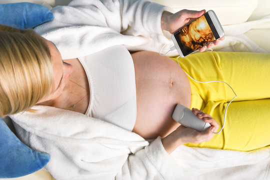Modern pregnant woman sitting on a white couch at home relaxing and doing by herself digital 3D ultrasound scan of a baby with a mobile smart phone
