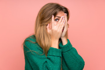 Young blonde woman with green sweater over pink wall covering eyes and looking through fingers