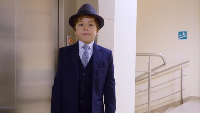 Kid boy looks like a businessman in suit and hat is waiting elevator, pressing the button and looking at camera in his office. Adult's life parody.