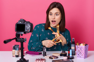 Shocked emotional young female records video about make up lifehacks, opening her mouth widely, holding various make up brushes in both hands, looks surprised. Beauty, style and blogger concept.