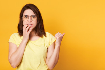 Frightened young attractive female holds her fingers on lips, shows aside with thumb, looks scared. Brunette emotional model wears yellow casual t shirt and spectacles. Copy space for advertisement.