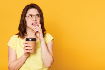 Confused thoughtful young lady looks aside, puts finger in her mouth, holds papercup with coffee in one hand, wearing light yellow casual t shirt and specstacles. Copy space for advertisement.