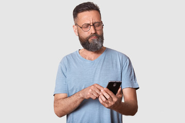 Serious calm brunette guy looks attentively aside, holding smart phone in his hand, typing messages to his friends, confused about posting. Handsome model wearing casual t shirt and fashionable specs.