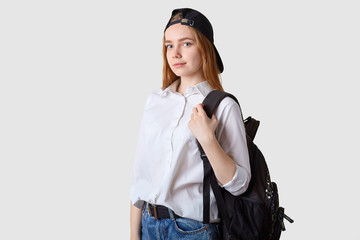 Slender beautiful energetic school girl goes to school wearing blue jeans, white shirt, black cap and bagpack, looks concentrated on studying, holidays come to end. Studying and education concept.