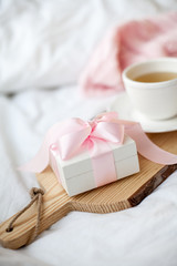 White mug with tea, gift box with ribbon on the bed. Breakfast in bed. Cozy. Pink plaid. Cotton.