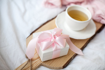 White mug with tea, gift box with ribbon on the bed. Breakfast in bed. Cozy. Pink plaid. Cotton.