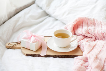 Fototapeta na wymiar White mug with tea, gift box with ribbon on the bed. Breakfast in bed. Cozy. Pink plaid. Cotton.