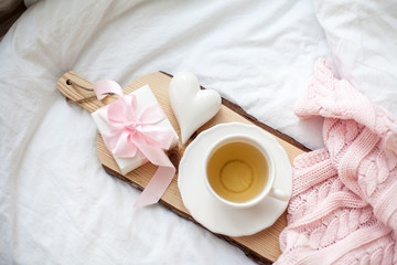 White mug with tea, gift box with ribbon on the bed. Breakfast in bed. Cozy. Pink plaid. Cotton.