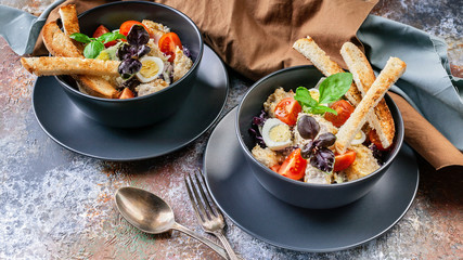 Two servings of Caesar salad with chicken, quail egg, cherry tomatoes, croutons and basil in dark blue bowls on a rusty background.