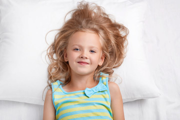 Attractive small kid with pleasant appearance, blue eyes and healthy skin looks at camera, poses in bed at bedroom, has refreshed look after having long sleep. Children and bed time concept.