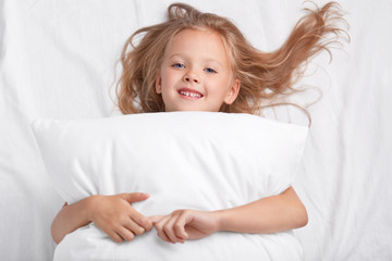Satisfied playful girl with charming smile, embraces pillow, lies on white pillow, has good rest, enjoys awakening, poses in bed. Children, rest, relaxation, lifestyle concept. Good morning.
