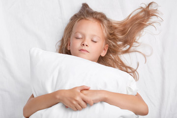 Obraz na płótnie Canvas Little attractive daughter with closed eyes, embraces soft white pillow during sleep, enjoys domestic cozy atmosphere, lies in white bed, being photographed by mother. Bedding and rest concept