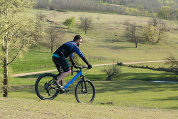 Cyclist in shorts and jersey on a modern carbon hardtail bike with an air suspension fork rides off-road on green hills near the forest