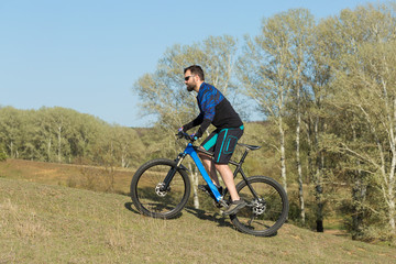 Obraz premium Cyclist in shorts and jersey on a modern carbon hardtail bike with an air suspension fork rides off-road on green hills near the forest