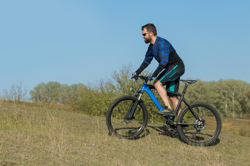 Fototapeta premium Cyclist in shorts and jersey on a modern carbon hardtail bike with an air suspension fork rides off-road on green hills near the forest