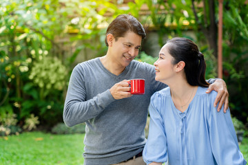 Asian senior couples are standing in the summer park and men are holding a red coffee mug. Which drinking black coffee during the day will make you feel refreshed. Life and health insurance concept